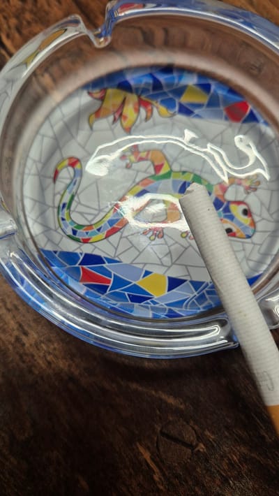 A close-up shot showcases a glass ashtray with a vibrant mosaic design of a colorful lizard. The lizard's body is depicted with geometric patterns in shades of green, red, yellow, and blue, set against a white tiled background. A single unlit cigarette rests diagonally across the ashtray, its white paper filter contrasting with the intricate artwork. The ashtray sits on a dark, textured wooden surface, creating a warm and natural backdrop.