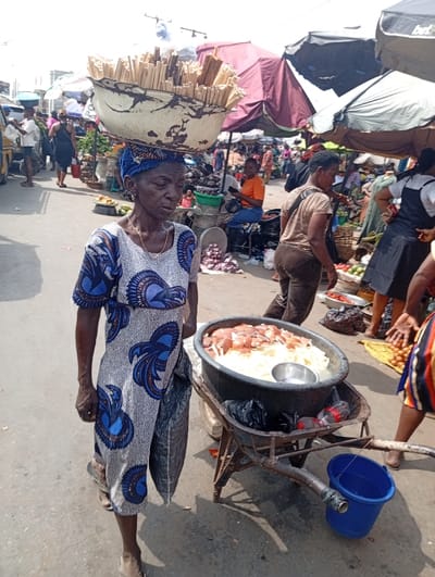 An elderly woman balances a large bowl filled with wooden sticks on her head, walking on a busy street market. She's wearing a patterned dress and carrying a black bag. A rusty wheelbarrow beside her holds a large container overflowing with white and reddish food items, and a small blue bucket hangs from the handle. The marketplace is bustling with people and covered with colorful vendor umbrellas.