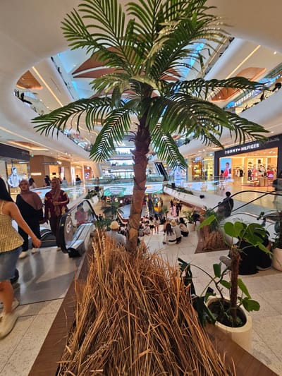 This image depicts the interior of a multi-level shopping mall in Denpasar, Indonesia. In the foreground, a decorative artificial palm tree stands prominently, surrounded by dried grass and potted plants, suggesting an indoor garden or decorative element within the mall. In the middle ground, escalators are visible, with several shoppers moving between levels. People are seen walking, standing, and interacting in various parts of the mall. Storefronts line the upper levels, with visible signage for brands such as "TOMMY HILFIGER" and "PEDRO," indicating a retail environment. The mall's architecture features modern design elements with curved white ceilings and wooden accents. The lighting appears to be a mix of ambient mall lighting and illuminated signage. The scene captures a typical day in a bustling urban shopping center.