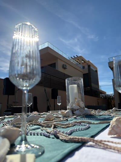 An outdoor table is set for a celebration with a teal tablecloth, decorated with seashells, rope, and white beaded strands. There are several champagne glasses on the table, one in the foreground is blurred and catches the sunlight. In the center of the table, a glass vase contains a white candle surrounded by shells. In the background, modern buildings with balconies and a bright blue sky with some wispy clouds are visible. Two black speakers are placed on stands to the left and right of the table. The location is described as being in Krimovice, Montenegro.
