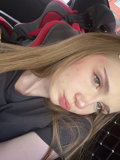 A young woman with light brown hair and green eyes is lying down, with her head propped up on her arm. She is wearing a dark grey t-shirt. Her hair falls around her face and shoulders. In the background, a portion of a red and grey car seat is visible. The scene appears to be inside a vehicle. There is no specific indication of the city of Sterlitamak, Russia within the image.