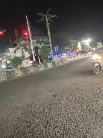 A night time street scene in Kutalhoksukon, Indonesia, is captured. In the background, a mosque with a blue domed roof is visible, adorned with intricate patterns. A traffic light, showing a red signal, is positioned on the left side of the frame. Palm trees and other greenery are scattered throughout the scene. A motorbike with its headlight on is in the foreground, suggesting movement and activity on the street. The road surface is asphalt, and there are concrete barriers along the side of the road. The overall atmosphere is illuminated by artificial lights, creating a nighttime urban environment.