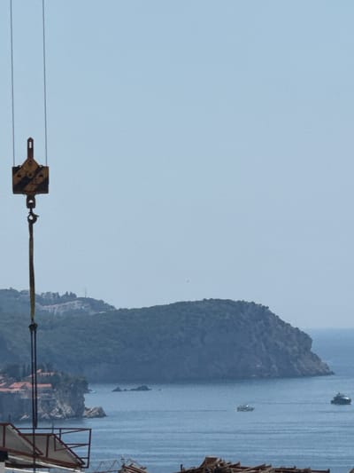 This image captures a scenic view of the Adriatic Sea near Boreti, Montenegro. A large, forested island dominates the right side of the frame, its steep slopes meeting the calm, blue water. Several boats are visible on the sea, one closer to the island and another further out. The sky is a pale, clear blue, suggesting a bright and sunny day, likely in the late morning or early afternoon given the quality of light.

In the foreground, a construction crane's hook and cable are prominently featured, indicating that some form of construction or repair work is in progress. The hook itself is yellow and black, with a sturdy metal construction. The cable is a thick, dark rope. These elements suggest a utilitarian aspect to the otherwise natural and picturesque scene.

The background shows a coastline with buildings nestled into the hillside, hinting at a coastal town or resort. The overall mood is one of tranquility and natural beauty, juxtaposed with the man-made industrial element of the crane. The clear weather and calm sea contribute to a peaceful atmosphere. No text is visible in the image.