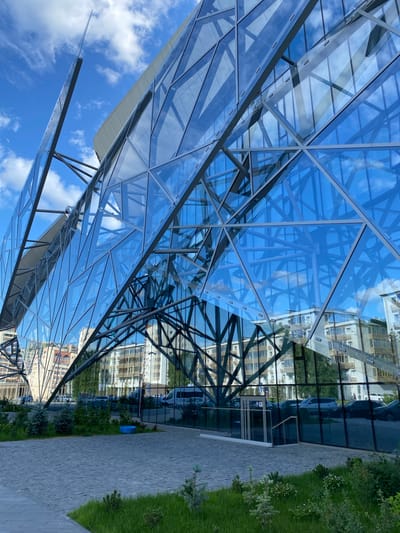 A modern, geometric building with a glass facade reflects the blue sky and white clouds. The building's structure is composed of triangular glass panels supported by a dark metal framework. The facade angles sharply upwards, creating a dynamic and futuristic appearance. In the foreground, a paved walkway leads to the entrance of the building, with green grass and small evergreen trees bordering the path. Beyond the glass, several multi-story residential buildings and parked cars are visible, suggesting an urban environment. The scene appears to be a public space in the city of Kazan, Russia, given the architectural style often found in contemporary developments there. There are no people prominently featured in the image, though a few figures are subtly visible in the reflections and in the background.