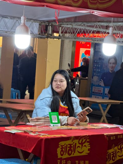An East Asian female is seated behind a red-draped table in an outdoor or semi-sheltered commercial area in Jiangjin, China. She wears a light blue puffer jacket over a red apron with yellow text and has long dark hair. Her gaze is directed slightly to her right. She holds a light-colored foldable smartphone in her left hand. On the table in front of her are stacks of paper items, possibly pamphlets or cards, and a green QR code sticker for mobile payment is prominently displayed. The red tablecloth features yellow text, including "利利心" and a smaller slogan below. Overhead, a metal truss structure supports a red fabric canopy and two large, brightly lit white LED light bulbs. In the background to the left, a partially obscured male figure in dark clothing stands near a blue stool. To the right, a large blue and white sign, identifying the location as "江津" (Jiangjin) and displaying "1300" and a female image, is visible. A person wearing a red helmet on a motorcycle or scooter is partially visible behind the woman to the right, and another individual in dark attire is also partially visible on the far right. Wooden tables with folding legs are present in the mid-background. The scene is illuminated by strong artificial light from the overhead bulbs, suggesting low natural light conditions.