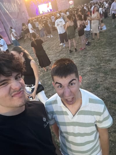 The image captures a crowd of people gathered outdoors, likely at a music festival, given the large stage and lighting visible in the background. The scene is set on a grassy area. Two young men are in the foreground, close to the camera. The man on the left is making a distorted facial expression, while the man on the right is shouting with his mouth open. In the middle ground and background, a diverse crowd is seen milling about, with some individuals dancing and others conversing. Several are holding mobile phones, suggesting they are taking photos or videos. In the far background, a large stage with vibrant purple and white lighting is prominent, with what appears to be performers on it, though they are not clearly discernible. A large banner or screen is also visible to the left, with text partially obscured. The overall atmosphere appears to be energetic and celebratory. The location is identified as Milano Music Week, Parco Musicisti in Segrate, Italy.