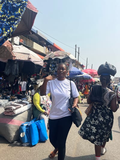 The image portrays a bustling open-air market street in Accra, Ghana, observed under bright daylight. In the foreground, a young adult female is walking forward, slightly angled left. She has short, dark hair, wears a white short-sleeved t-shirt, black leggings, and light brown sandals. Her left hand is raised to the level of her head, palm open, while her right hand holds a black smartphone and the strap of a black tote bag carried on her left shoulder. Parallel to her, on the right, an adult female is walking away from the camera. This individual carries a large black plastic bag balanced on her head, wears a patterned sleeveless top, a dark skirt with white floral patterns, white socks, and dark sandals, and holds a dark, brush-like object in her right hand.

The background features numerous market stalls lining the street, many shaded by diverse sun umbrellas in colors including white, blue, red, and brown. Merchandise visible includes rolls of fabric, patterned textiles, and various clothing items hung on displays. A vendor in a bright yellow long-sleeved top is seated behind a stall on the left, near a display of small packaged goods. Behind the stalls, multi-story commercial buildings with balconies and red and blue architectural elements are visible. Overhead utility poles and wires traverse the scene against a clear, pale blue sky. The ground is an asphalt surface. Additional pedestrians and vehicles are discernible further down the crowded street.