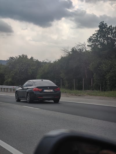 The image shows a black BMW 4 Series Gran Coupé driving on a highway in Busmantsi, Bulgaria. The car is in the right lane, and the perspective is from another vehicle driving behind and to the left. The license plate of the BMW is visible and reads "D-SONMEZ".

The scene is an overcast day, with a cloudy sky. Dense green trees line the sides of the highway, with a few bare branches visible amongst them. A metal guardrail separates the highway from the roadside. In the foreground, the mirror of the vehicle taking the photo is visible, reflecting a blurred image. The overall impression is of a typical highway drive on a cloudy day.