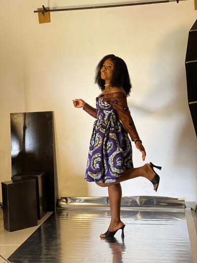 A young Black woman stands in a studio setting. She is wearing a short, off-the-shoulder dress with a blue and white swirling pattern. The dress has short, puffy sleeves made of brown fabric. She is also wearing black heeled sandals and a pearl necklace. The woman is posing with one leg raised behind her. The background is a white wall, with a black rectangular object to the left of the woman. The floor is covered in a reflective silver material, with a black and white tiled section visible in the foreground. There is a black photography light on the right side of the frame, partially visible. The location is described as Durumi, Nigeria.
