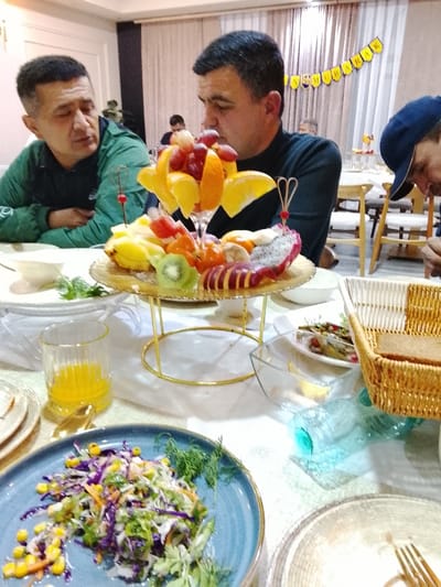 A table setting features a lavish fruit display with oranges, grapes, kiwi, and dragon fruit on a tiered golden stand. Two men are seated nearby, one in a green jacket and the other in a black turtleneck. Additional dishes include a salad on a blue plate and a bread basket. A glass of orange juice is visible, along with other table settings, suggesting a celebratory gathering. A banner is hanging in the background.