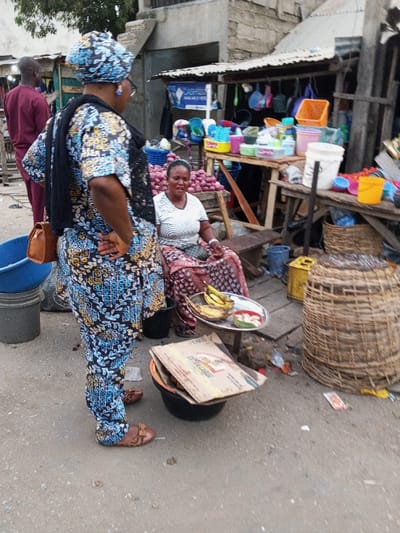 A woman in a patterned blue outfit speaks to a seated vendor. The vendor sits before a display of bananas and a basket, possibly selling fruit. Behind them, another stall displays colorful plastic items and onions. The scene seems to be a bustling outdoor market, with a man in a maroon shirt walking in the background. The area appears to be a bit cluttered and dusty.
