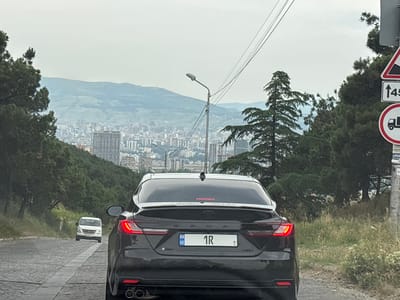 The image shows the rear of a black Toyota Camry driving on a cobblestone road in Tbilisi, Georgia. The car has a Georgian license plate that reads "GE 1R". In the background, a cityscape with numerous buildings is visible, situated on a hillside and extending into the distance. Mountains can be seen beyond the city. A white minivan is in front of the Camry, and a traffic sign indicating a steep descent is visible on the right side of the frame. The sky is overcast.