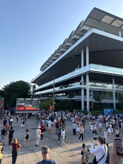 The image shows a crowd of people gathered outside a modern building under a clear blue sky. The building has a sleek, angular design with visible architectural elements. A large digital scoreboard displays information, possibly related to a sporting event. The crowd appears to be diverse, with people of various ages and ethnicities. Some individuals are holding up their phones, possibly taking pictures or videos. The setting seems to be a public space, likely an entrance or plaza associated with the building.
Based on the image's elements, this is Roland Garros in Paris, France. The scoreboard shows the results of tennis matches, and the crowd likely consists of spectators attending the tournament.