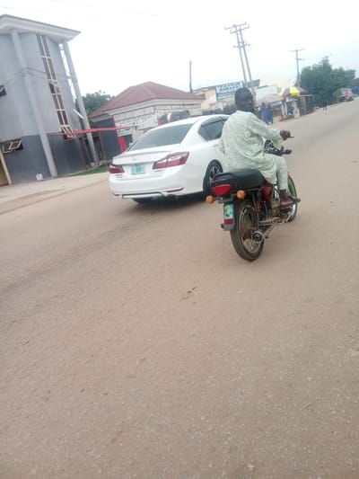 A medium shot captures a man riding a red and black motorcycle on a paved road in Kubwa, Nigeria. The man is wearing a light green patterned tunic and pants, and he is looking towards the camera. He is positioned on the right side of the image, with the motorcycle's rear wheel in the foreground. To the left of the motorcycle, a white Honda Accord sedan is parked or moving slowly. The car's license plate reads "ABC 694K". In the background, several buildings are visible, including a light gray structure with pillars on the far left and a building with a reddish-brown roof further back. A blue sign with white text that reads "FAVOUR EMBASSY" is also visible in the background. The sky is overcast, and the road appears to be dusty. The scene suggests a typical street view in an urban or semi-urban area of Nigeria.
