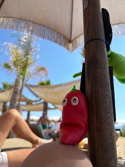 The image, taken at Ofrynio Beach, Greece, shows a close-up of a red, toy pepper with wide, cartoonish eyes, attached to a wooden umbrella pole. The toy is positioned in the foreground, obscuring part of the viewer's body which is resting on a lounge chair. In the background, palm trees and white fabric sunshades are visible, with the bright blue sky overhead. Out of focus, two individuals can be seen lounging in the background. A green toy is also attached to the umbrella pole, to the right of the pepper toy.