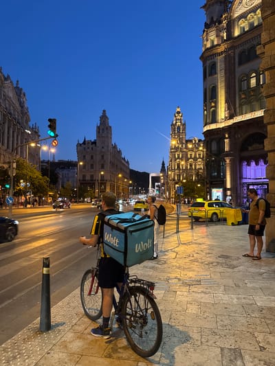 The image shows a person on a bicycle, wearing a blue "Wolt" delivery bag on their back. The person is looking towards the right, with their hands on the handlebars. The bicycle has a black frame and a black rack on the back. The person is wearing a yellow and black shirt and dark shorts. They are wearing athletic shoes.
The scene is an urban street at dusk, with buildings on either side of the road. The buildings are lit up, suggesting it is evening or night. The sky is a deep blue, indicating twilight. There is a yellow taxi visible in the background. A traffic light is also visible, showing a green light. The street is paved with cobblestones. There are other people visible in the background, some walking and some sitting.
The overall mood of the image is one of urban activity and a sense of movement. The time of day and the illuminated buildings create a vibrant atmosphere. The presence of the delivery bag suggests that a delivery service is operating. The location appears to be a city street in Budapest, Hungary, based on the provided context. There is visible text on the blue bag that says "Wolt" in white letters. There is also text on the yellow taxi, but it is not clearly legible.