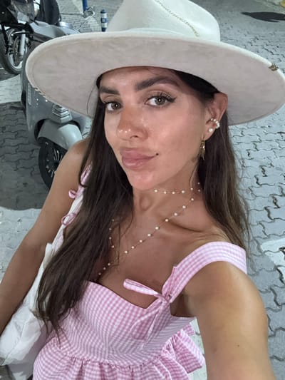 A woman wearing a wide-brimmed hat and a pink and white gingham dress is captured in a selfie. She has long dark hair, brown eyes, and is wearing pearl earrings and a pearl necklace. Her lips are slightly parted. The background shows a cobblestone ground, part of a scooter, and what appears to be a street in Fethiye, Turkey, possibly near a harbor or waterfront area, suggested by the scooter and paving stones.