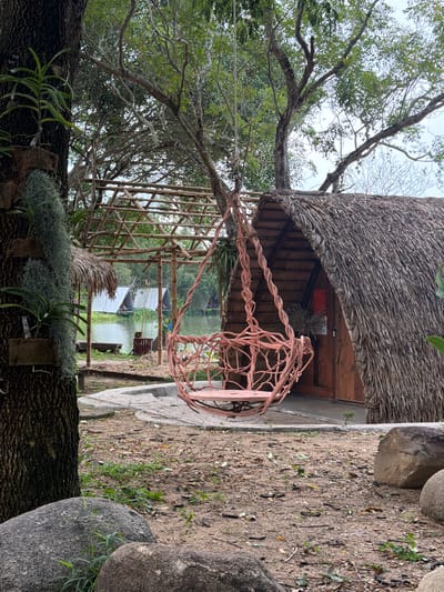 The image depicts an outdoor scene in Khánh Vĩnh, Vietnam, characterized by a rustic, natural setting. In the foreground, a large tree trunk with textured bark is prominent on the left, adorned with hanging plants and epiphytes. A pink, woven swing chair hangs from a tree branch, positioned slightly to the right of center. Behind the swing, a small, A-frame hut with a thatched roof and wooden door is visible. To the left of the hut, a bamboo structure with a thatched roof extends over a path leading towards a body of water. In the background, the water is dotted with several white tent-like structures on rafts or boats, suggesting a resort or accommodation area. The ground is covered with dirt, fallen leaves, and some scattered stones, indicating a natural, unpaved area. Lush green foliage and trees frame the scene, contributing to the serene, natural ambiance.