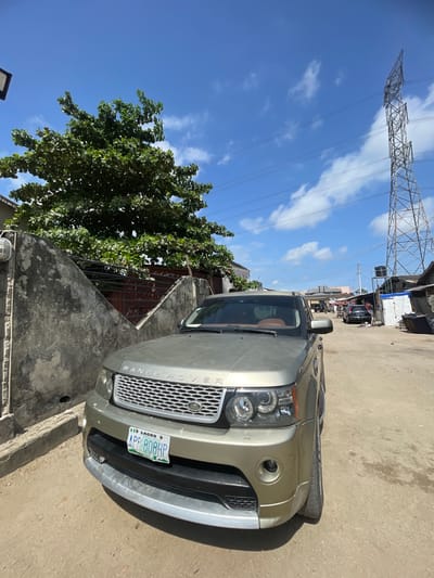 The image shows a gold Range Rover parked on a street. The license plate reads "APP 808HP" and indicates it is registered in Lagos, Nigeria. The car is partially obstructed by a concrete wall and a tree. In the background, there is an electricity pylon and other buildings lining the street. The sky is blue with scattered clouds. While there is no Ado, Nigeria. There is Ado Ekiti, Nigeria. There is no information in the image to verify the image's location.