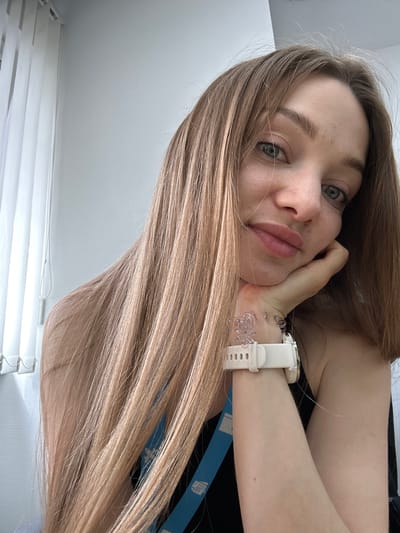 The image is a bright, close-up selfie of a young woman, taken indoors in Kazan, Russia. She has long, straight, light brown or ash blonde hair that cascades across the left side of the frame. Her eyes are a striking light green or hazel, and she looks directly at the camera. Her face shows a relaxed expression with full, slightly pursed lips in a subtle pout, and she rests her chin gently on her right hand.

She is wearing a dark, possibly black, top. On her left wrist, she wears a white Garmin smartwatch, with the brand name "GARMIN" distinctly visible on the strap. Below the watch, she has a decorative clear bracelet, possibly made of plastic or silicone, featuring interconnected floral or butterfly-like shapes.

The background is minimalist, consisting of plain, light-colored walls. White horizontal window blinds are visible in the lower left and right portions of the image, suggesting the presence of a window and indicating that the photo was taken during the daytime with ample natural light. The overall mood is casual and well-lit.