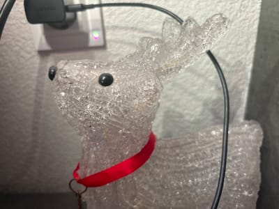The image captures a close-up of a translucent decorative reindeer statue positioned indoors. The reindeer is crafted from a clear, textured material, giving it a crystalline or icy appearance, and features small black bead eyes. It wears a bright red ribbon around its neck, from which a small metal ring hangs.
In the blurred background, a black "SAMSUNG" branded power adapter is plugged into a white electrical outlet mounted on a light-colored, textured wall. A black cable extends from the adapter, running diagonally across the upper portion of the frame, partially obscuring the reindeer's antlers. The power outlet displays a small, glowing indicator light with green and pink hues, suggesting it is active.
The scene is static, depicting a decorative item in a home or similar indoor environment. No explicit activity or event is taking place. The visible text includes "SAMSUNG" on the power adapter. The location context for this media file is Kuklen, Bulgaria.