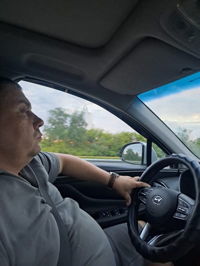 The image depicts the interior of a car, focusing on the driver from a passenger's perspective. The original image is rotated 90 degrees counter-clockwise. A person, presumably male, is seated in the driver's seat, wearing a gray t-shirt and a seatbelt across their chest. A brown leather-strapped watch is visible on their left wrist, which rests on the steering wheel. The driver's face is seen in profile, looking forward and slightly upwards, with a neutral expression.
The steering wheel, featuring a black cover for grip, has a prominent Hyundai logo in its center, indicating the car's make. Various control buttons are visible on the steering wheel spokes. Through the car windows, a natural landscape of green trees and an overcast or partially cloudy sky can be seen, suggesting the vehicle is in motion on a road or highway. The driver's side rearview mirror reflects the passing scenery. The car's interior, including the dashboard and overhead lining, appears standard.
The activity taking place is driving. The time of day is ambiguous due to the cloudy sky, but it is daylight. No specific location cues beyond the natural landscape are visible to pinpoint the exact location within Mayak, Russia. No other significant text apart from the car's emblem is discernible.