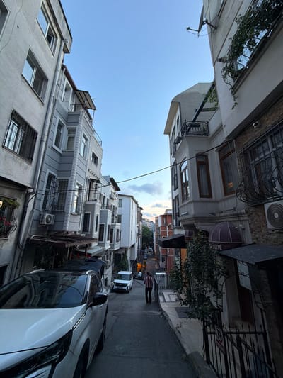 This is a street-level, slightly upward-angled, full-shot photograph of a narrow, uphill street in Beyoğlu, Turkey, at dusk. The sky is a clear, light blue with some wispy clouds on the horizon. The street is flanked by multi-story buildings with ornate architectural details, characteristic of the area.

On the left, a light gray building dominates the foreground, featuring several windows with metal grates and air conditioning units. Further up the street, a similar building with light gray facades and white trim is visible. On the right, a building with a brown brick facade is in the foreground, with a white stucco building next to it. This building has large, bay windows with wooden frames and decorative ironwork on the upper floors. A black awning with the word "SUITE" is visible above the entrance of a business.

In the street, a white SUV is parked in the foreground with its trunk open. Further up the street, a white minivan is parked, and another car is visible in the distance. A single person, wearing a red and white striped shirt and dark pants, is walking uphill away from the camera. The street is paved with asphalt, and there are sidewalks on both sides. Trees and shrubbery are visible, adding greenery to the urban landscape. Power lines are strung across the street overhead. The overall impression is of a residential or commercial street in an older European-style city.