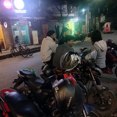 A group of four young men are gathered on motorcycles on a street at night. They appear to be in casual conversation. The motorcycles are parked near a brightly lit shop, "Kalamati Anna." A couple of bicycles are parked nearby. Other vehicles are also visible further down the road. Several helmets are placed on the motorcycles, and the atmosphere seems relaxed and friendly.
