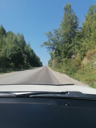 The image is taken from the perspective of someone inside a vehicle, looking forward through the windshield. The vehicle is on a paved road that curves slightly to the right and appears to be going uphill. The road is bordered on the left by a steep, tree-covered slope and on the right by a less steep, wooded hillside. The trees are predominantly green and appear to be deciduous, with some coniferous trees mixed in. The sky is clear and blue. In the distance, on the right side of the road, there are two white cars. The foreground of the image shows the interior of the car, with the dashboard and windshield wipers visible. The location is identified as the city of Uşanovo, Kazakhstan.