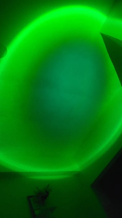 This image depicts an interior space in Nanyuki, Kenya, characterized by a dominant circular green light projection. The light, brighter at its periphery, illuminates a wall or ceiling surface, creating a gradient that darkens towards its center. Architectural features are discernible: a prominent corner where two walls meet extends diagonally downwards from the upper right, and another wall intersection is visible in the upper left. A horizontal line in the lower central area may indicate a wall-ceiling junction or a shelf. In the lower-left quadrant, a small, indistinct, dark-toned object, possibly a decorative item or plant, rests on a surface, rendered in silhouette by the green illumination. The overall scene is devoid of individuals, active subjects, or discernible interactions.