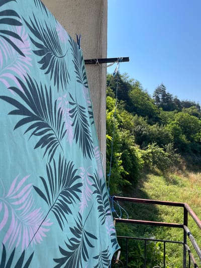 A light blue beach towel with a dark gray and light pink palm leaf pattern is hung to dry on a clothesline attached to the side of a concrete building. The beach towel is secured with blue clothespins. A dark metal railing is visible to the right of the towel, with a green, grassy hillside and trees in the background. The sky is clear and blue. The scene appears to be on a balcony or porch, possibly in Zlatograd, Bulgaria, given the mountainous terrain and vegetation.