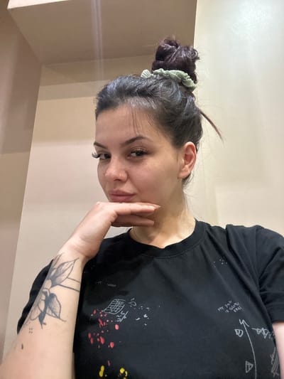 The image features a young woman posing indoors against a plain, light-colored wall. Her head is tilted, and she looks towards the viewer with a calm, slightly smirking expression, her left hand resting on her chin. She has dark hair tied in a bun with a light green scrunchie, noticeable long eyelashes, and visible freckles on her face.

She wears a black t-shirt adorned with white geometric patterns, red and white paint splatters, and vertical text that reads, "MY [partially obscured word] IS GLUTEN FREE". A black ink tattoo depicting a swallow or similar bird with floral elements is visible on her right forearm, accompanied by small dot tattoos. The scene captures a casual indoor moment, set in Sliven, Bulgaria. The lighting appears natural for an indoor setting, and no specific time of day is discernible.