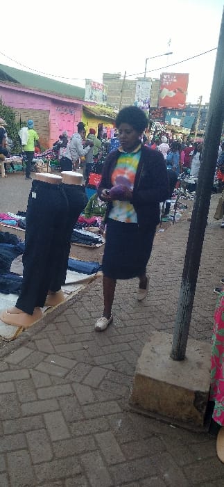 An outdoor scene shows a bustling market with clothes displayed. A woman wearing a tie-dye shirt, a black jacket, and a dark skirt appears to be browsing. The clothing is laid out on what appears to be a white sheet on the ground. Paved brick makes up the ground. A pole stands upright with its base cemented into the ground. Buildings and more people are visible in the background, adding to the lively atmosphere of the marketplace.
