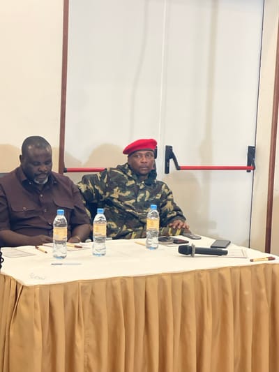 An indoor photograph depicts two adult males seated at a table in Nyayumba, Rwanda. The man on the left, with dark skin and a dark beard, wears a brown button-down shirt and looks downwards. The man on the right, also with dark skin, wears a camouflage jacket and a red beret, looking forward. Both are seated behind a table draped with a white tablecloth over a beige pleated skirt. On the table are three clear plastic water bottles with yellow labels, multiple pens (brown, blue, yellow), at least three smartphones, and a black microphone. The background consists of a plain white wall with a vertical brown trim on the left and a red horizontal emergency exit push bar assembly on the right.