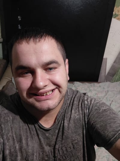 The image shows a close-up selfie of a man with fair skin, short dark hair, and blue eyes. He is smiling broadly, revealing some of his teeth. He is wearing a dark grey t-shirt with a textured pattern. The background appears to be a dark surface, possibly a wall or furniture, with some patterned fabric visible on the right side. There are also some faint lines and shapes on the background that could be part of a poster or wallpaper. The lighting suggests an indoor setting, possibly at night or in a dimly lit room. The man is positioned slightly off-center, with his head tilted towards the camera. The overall impression is of a casual, personal photograph taken by the subject himself. The location of Ketrysanivka, Ukraine is not visually identifiable within the provided image.