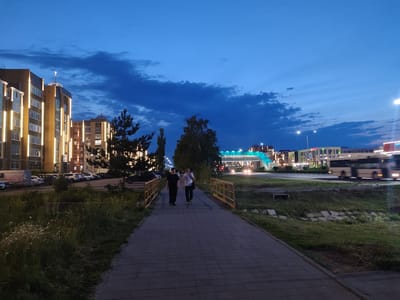 The image captures a twilight scene in Kostanay, Kazakhstan, likely near the "Shanyrak" residential complex. Two individuals are walking on a paved walkway. The person on the left is wearing a dark t-shirt and pants, while the person on the right is in a white t-shirt and dark pants, carrying a bag. They are walking away from the viewer, towards a bridge with yellow railings.
On the left side of the walkway, there is a patch of grass with wildflowers and a line of parked cars. Behind the cars, a modern apartment building is visible, illuminated by vertical lights. In the background, across a road, another building with extensive lighting and a pedestrian overpass with blue and green elements is prominent. A bus is moving on the road in the middle ground. The sky is a deep blue with scattered clouds, indicating the transition from day to night. The environment appears to be a developing urban area with residential and commercial buildings.