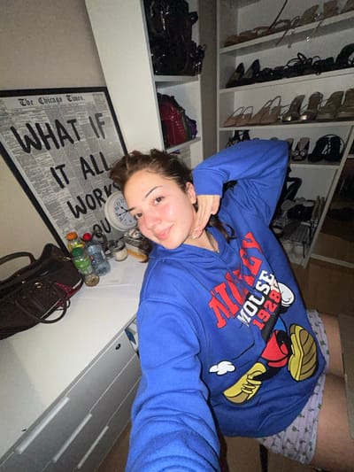 A selfie of a young woman wearing a blue Mickey Mouse hoodie. She's lying down with her head near a floor mat with a newspaper print that reads "What If It All Works Out." A clock, water bottles, and a handbag are scattered near the mat. Behind her are shelves filled with shoes and some handbags. The background includes a white storage unit.