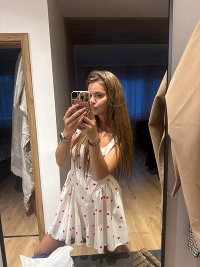 A young woman is captured in a mirror selfie within an indoor setting, identified as Plovdiv, Bulgaria. She stands in a room with light-toned wooden flooring, wearing a white, sleeveless dress adorned with numerous small red heart patterns. Her long, straight brown hair extends down her back and over her shoulders. On her left wrist, she wears a silver-colored smart watch, and on her right, a dark-colored bracelet. Her fingernails are painted pink.

She holds a smartphone with a decorative light-colored case featuring a pink circular design and celestial patterns, positioned in front of the lower half of her face. Her lips are slightly pursed, and her head is tilted. The mirror reflects a segment of a white wall on the left, adjacent to a wooden door frame, which reflects a light-colored garment draped behind it. To the right, a beige garment hangs on a dark rack, with a small portion of a black-and-white checkered pattern visible beneath it. In the background reflection, a darker interior space is visible through an opening, featuring what appears to be dark blue vertical blinds or curtains. On the floor in the foreground, a white shopping bag with blue accents and a brown patterned bag are present.