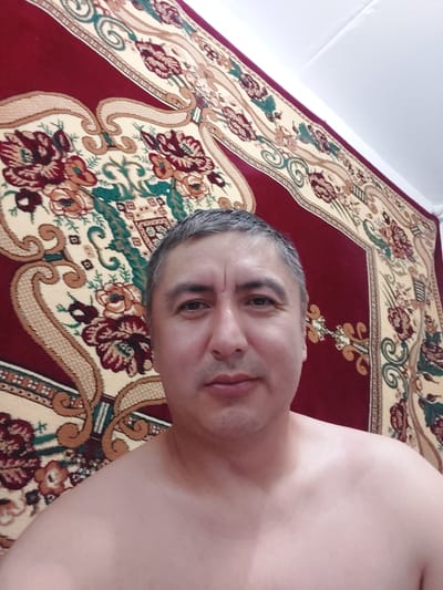 A man with graying hair is pictured from the chest up, looking directly at the camera. He is shirtless, and his face is in sharp focus. The background consists of a large, ornate rug with a maroon border and a cream-colored field featuring floral and scrollwork patterns in shades of red, green, and gold. The lighting suggests an indoor setting, likely a room in a residential building. The image does not provide specific geographical information related to the city of Ması, Kyrgyzstan.