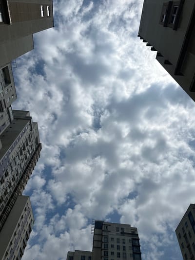 The image is taken from a low angle, looking up at the sky framed by several buildings. The sky is filled with scattered, greyish-white clouds, with patches of blue sky visible in between. The buildings are tall, residential apartment blocks, common in densely populated urban areas. They are made of concrete and have numerous windows. The architectural style is modern but unremarkable. There are no visible people, vehicles, or street-level details, focusing instead on the sky and the upper parts of the buildings. The image composition suggests a confined urban space, typical of high-density living. The location is potentially Esenyurt, Turkey, based on the prevalence of similar architecture in that area.