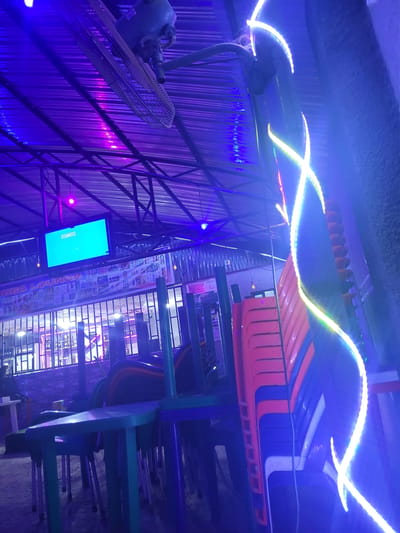 The image depicts a dimly lit outdoor seating area of what appears to be a lounge or restaurant in Madalla, Nigeria. The scene is illuminated by vibrant purple and blue neon lights, creating a nocturnal ambiance. A large industrial fan is mounted on the ceiling, directing air downwards. In the background, a television screen displays a bright turquoise image. Stacks of chairs, primarily orange and blue, are visible, suggesting the area may be closed or preparing for patrons. The ground is covered in sand or gravel. A sign that reads "VIBES LOUNGE" is partially visible above a metal-grated barrier. The overall impression is one of a commercial establishment setting up for evening service or winding down after a busy period.