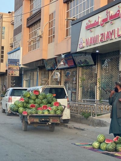 The image shows a street scene in Kandahar, Afghanistan. A small cart is piled high with watermelons, some cut open to display the red flesh. The cart is parked on the side of the road next to a pile of watermelons on a colorful cloth. Behind the cart are two light-colored vans. A man in traditional Afghan clothing, including a pakol hat and a vest, stands next to the watermelons on the ground and appears to be talking on a phone. The buildings in the background are multi-story with a mix of windows and closed metal security gates. A sign in Arabic script above a store reads "NAWI ZIYAYI LTD". An air conditioning unit is attached to the building. The light suggests it is either early morning or late afternoon.