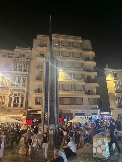 The image depicts a lively night scene in the city of Málaga, Spain, specifically on a wide pedestrian street illuminated by artificial lights. A tall, modern-looking monument, appearing to be made of concrete and dark tiles, stands prominently in the center of the frame, dividing the crowd. Surrounding the monument, a diverse group of people are gathered, walking, standing, and socializing.

On the left side of the street, a multi-story building with ornate architecture, featuring arched windows and decorative elements, is visible. To its right, another building with a more contemporary design, characterized by a series of balconies and uniformly shuttered windows, rises up. Further to the right, a yellow-painted building with bay windows adds to the urban landscape.

Several establishments are identifiable, including an "IRISH PUB" with a Guinness sign, and another place advertising "MORRISSEY'S". Other signs suggest businesses such as a "TASQUITA DE EN MEDIO" and a cannabis shop. The presence of tables and umbrellas indicates outdoor seating for patrons.

People in the image are dressed in casual summer attire, ranging from dresses and skirts to shorts and t-shirts. They are engaged in various activities, with some appearing to be moving through the area, while others are in small groups, conversing. The atmosphere suggests a vibrant social gathering and a bustling nightlife.

In the foreground, a few individuals are captured in more detail. A woman in a floral dress walks towards the viewer, accompanied by another woman in white pants. A man in a white t-shirt and shorts is also visible, as well as another man in a black t-shirt and shorts. A sculptural element, possibly a bronze statue of a person, is partially visible on the left side of the frame.

The ground is paved, and some litter is scattered in places, indicating the busy nature of the location. The overall scene is characterized by a mixture of architectural styles, commercial activity, and a significant human presence enjoying the night.