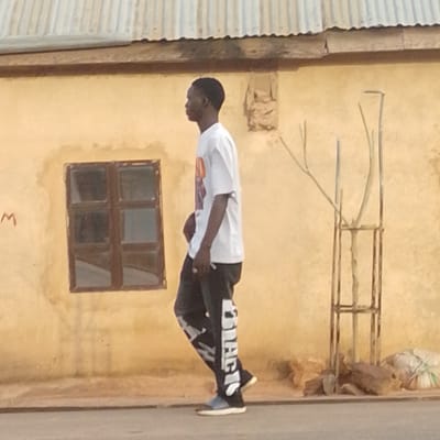 A young Black male is walking in profile to the left, wearing a white t-shirt with an orange and white graphic on the chest, and dark pants with large white lettering down the sides. He is wearing blue and white sneakers. He is walking past a light yellow building with a corrugated metal roof. To the left of the male is a small, dark-framed window with six panes. To the right of the male is a metal structure, possibly a trellis or support for a plant, with some bare branches. There are rocks and a white plastic-wrapped object on the ground to the right of the metal structure. A faint red letter "M" is visible on the building to the left of the window. The scene appears to be a street in Tempure, Nigeria.