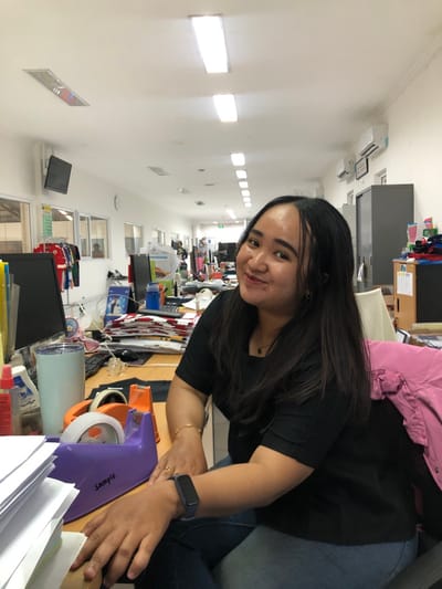 This image depicts an indoor office environment in Semarang, Indonesia. In the foreground, a woman with long, dark hair, wearing a black short-sleeved top and blue jeans, is positioned, looking directly at the camera with a slight smile. She has a dark smartwatch on her left wrist and a gold-toned bracelet on her right. Her hands rest on a desk surface. The desk features a stack of white papers, a light blue insulated cup, a bottle of hand sanitizer, a purple tape dispenser labeled "Sample," and a portion of a black computer monitor. A pink jacket is draped over a chair to the woman's right. The background shows a long, brightly lit office corridor. The white ceiling contains numerous recessed fluorescent lights, and air conditioning units are mounted on the right wall, near a dark grey filing cabinet. Multiple workstations with computers and documents are visible along the corridor. Through a windowed partition on the left, racks displaying red and white garments and a partial silhouette of another individual are discernible. An illuminated "Exit" sign is visible further down the hallway.
