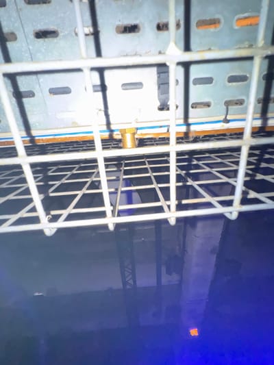 The scene presents an inverted, upward-looking perspective captured through a white metal wire grid, obscuring the immediate foreground. Beyond the grid, a perforated grey metal cable tray is visible, featuring horizontal rectangular slots and a brass-colored threaded fitting. A narrow blue and white stripe runs along the base of this tray, which exhibits minor dust and debris accumulation. Below the grid, a highly reflective, dark blue floor dominates the lower two-thirds of the image, mirroring overhead industrial structures. Reflected elements on the floor include a dark, angular metal truss structure on the left, a large rectangular support beam towards the right, and a small, distinct orange rectangular light source or object reflected in the far bottom-right. The environment is dimly lit by artificial light, contributing to the blue and purple tones of the reflective surfaces. This setup is consistent with a utilitarian interior space, such as a service tunnel, technical basement, or equipment room within a major infrastructure facility or large commercial building in Paris, France. No people, subjects, actions, or interactions are discernible.