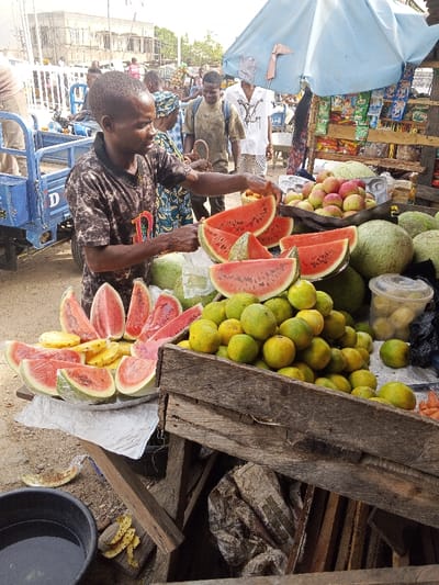This image captures an outdoor fruit stall operating in an urban street market environment within Zango-ndaji, Nigeria. In the foreground, a male vendor, wearing a dark patterned t-shirt, stands behind a display of fresh fruit, actively arranging slices of watermelon. His left hand is on a piece of watermelon, while his right hand hovers near another.

The stall features several platters holding numerous pre-cut watermelon slices, some individually wrapped in clear plastic. Adjacent to these are wrapped slices of pineapple. To the vendor's right, a large pile of green-yellow citrus fruits (likely oranges) fills a wooden crate, with more scattered on the ground. Behind these, several whole watermelons are visible. A metal scale containing red apples is positioned further back on the stall. A clear plastic container holding yellow fruit is also present. Above the fruit display, a light blue market umbrella provides shade, and a small shelf underneath it holds packaged snack items.

The ground around the stall is dirt, with scattered fruit peels (watermelon and pineapple) and a black plastic basin filled with water visible to the left. In the mid-ground and background, multiple individuals are present, suggesting a bustling market scene. A blue motorized tricycle (cargo variant) is partially visible on the far left. Buildings, some appearing to be under construction, form the backdrop.