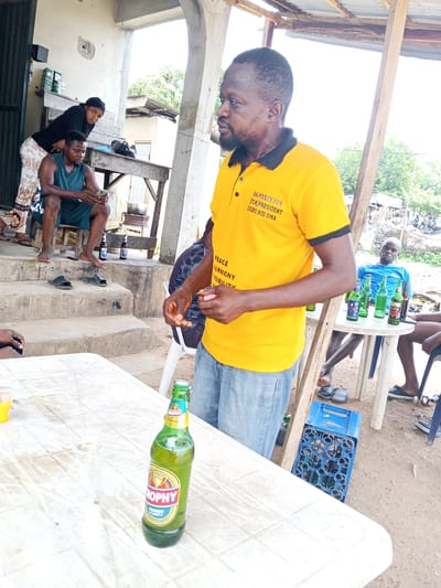 The image shows a group of people gathered in what appears to be an outdoor social setting, possibly in Zango-ndaji, Nigeria, based on the context. A man in a yellow polo shirt with political messaging is standing prominently in the foreground. On a white table in front of him, there is a bottle of Trophy lager beer. To the left, there are individuals sitting and standing under a veranda. A man is seated on a stool, looking at a phone. A woman stands behind him, looking at the camera. Further to the right, another man is seated at a table with several beer bottles. A blue plastic crate sits on the ground beside the table. The environment appears to be a casual, outdoor space with a mix of building structures and natural elements.