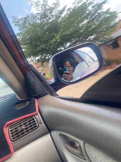This is a first-person perspective image taken from inside a car, likely a sedan, with the camera held by a person. The person is reflected in the side-view mirror, holding a smartphone and taking a selfie. They have dark skin, braided hair, and are wearing a dark shirt. The reflection also shows the interior of the car and the scenery outside.

The exterior scene visible in the mirror and through the car window includes a tall tree with dense green foliage, a brick building in the background, and a cloudy sky. There are also hints of other vehicles, including a motorcycle parked on the street. The lighting suggests it is daytime, possibly late morning or early afternoon, with bright sunlight filtering through the clouds.

The interior of the car is partially visible. The dashboard is a neutral beige color. A black speaker grill is visible near the side mirror, and a floor mat with a pink trim is on the floor. A vent with adjustable slats is also prominent on the dashboard. The overall impression is of a casual, everyday moment captured while in transit or stopped in a residential or semi-urban area. The setting appears to be Gura Top, Nigeria, as indicated by the location context. No visible text is present in the image.