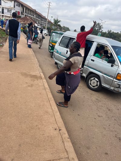 In the city of Gura Top, Nigeria, a group of individuals are gathered on a street. The scene is characterized by a concrete sidewalk and a paved road, with buildings lining the background. A man in a black vest and blue jeans walks on the sidewalk, facing away from the camera.

On the road, a silver van with its doors open is a central focus. A man wearing a red shirt stands partially inside the van, with his arm raised. Next to the van, a woman in a tan and black shirt and purple pants is positioned with her back to the camera, appearing to be interacting with someone within the vehicle. A woman in a green shirt is seated inside the van, looking at her phone.

Several other vehicles, including a green and white minivan and a motorcycle, are visible on the road, suggesting a busy street environment. The sky is overcast. The overall impression is one of everyday street life and transportation in a Nigerian urban setting.